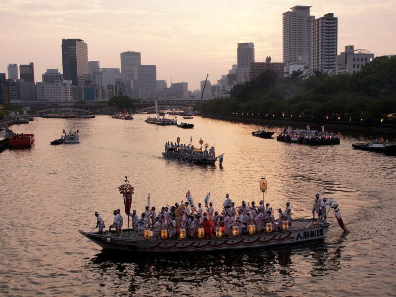 Lantern-lined festival boats carry participants along Osaka's Okawa River, with bridges and city buildings behind them at dusk.