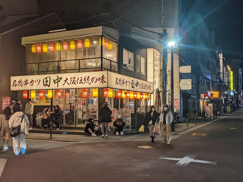 Lanterns illuminate a corner restaurant as pedestrians gather on Osaka's Amerikamura streets at night.