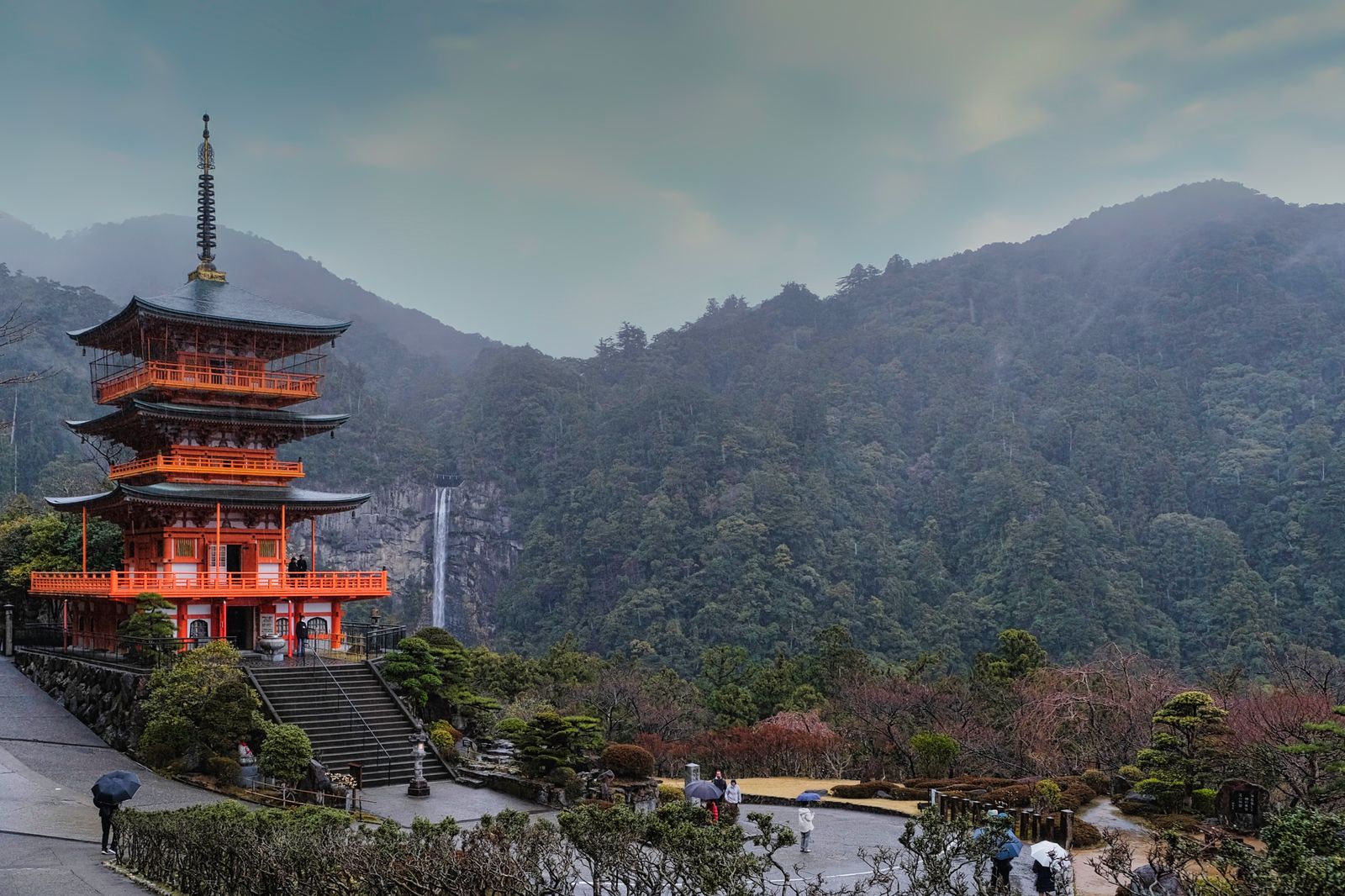 Visitors with umbrellas below the red pagoda of Seiganto-ji, with Nachi Falls in the misty hills behind
