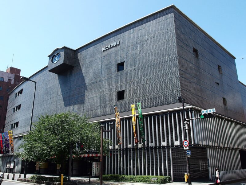 National Bunraku Theatre in Osaka with a projecting clock, grey facade and colourful banners beside the entrance.