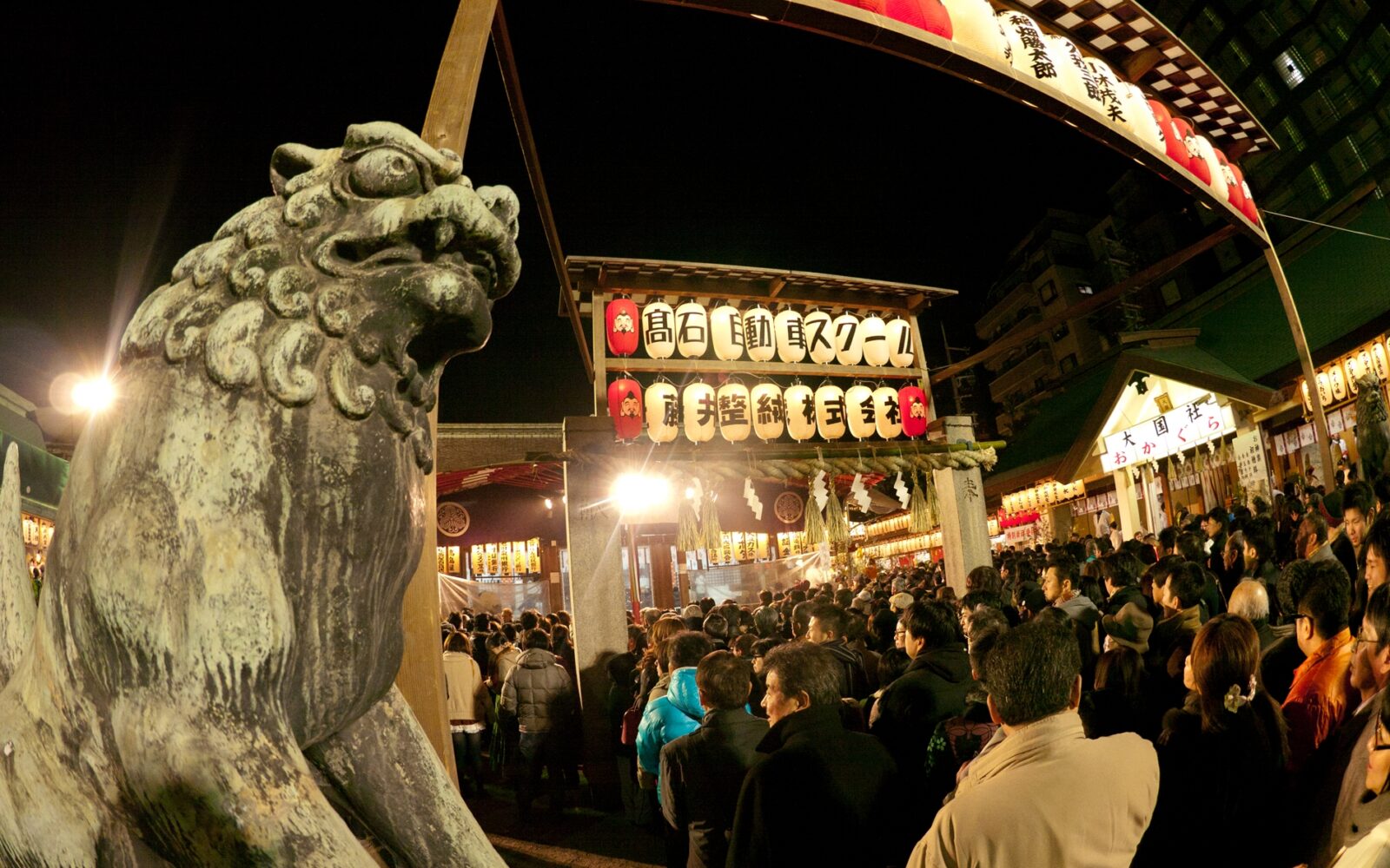 Nighttime visitors crowd Imamiya Ebisu Shrine in Osaka beneath rows of lanterns, beside a stone guardian lion.