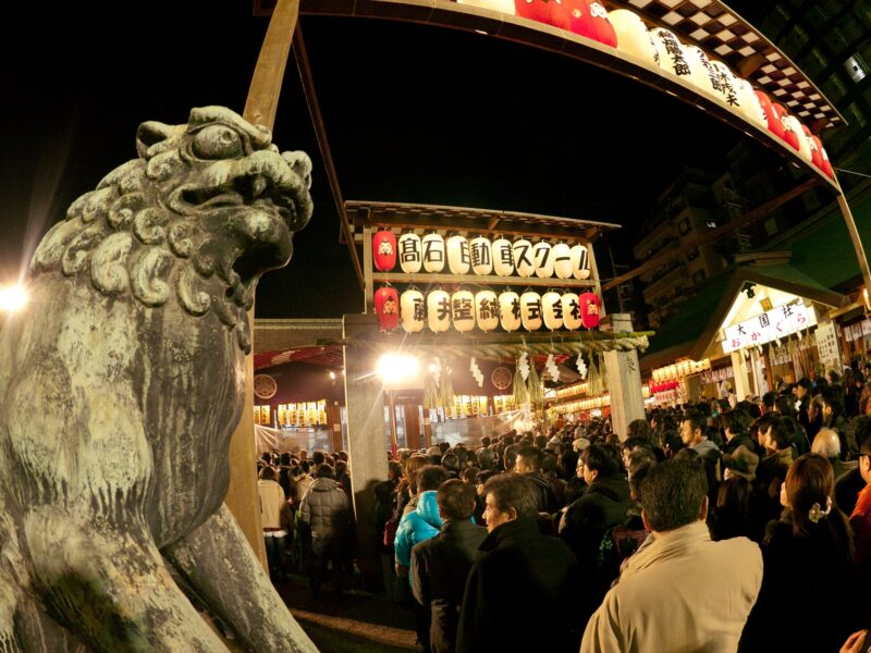 Nighttime visitors crowd Imamiya Ebisu Shrine in Osaka beneath rows of lanterns, beside a stone guardian lion.