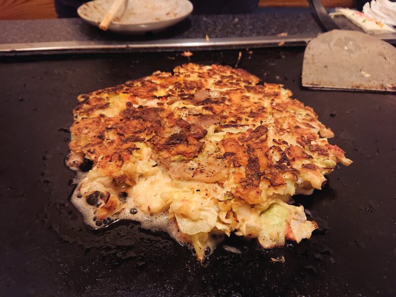 Okonomiyaki cooks on a restaurant griddle in Namba, Osaka, with browned cabbage batter and a metal spatula beside it.