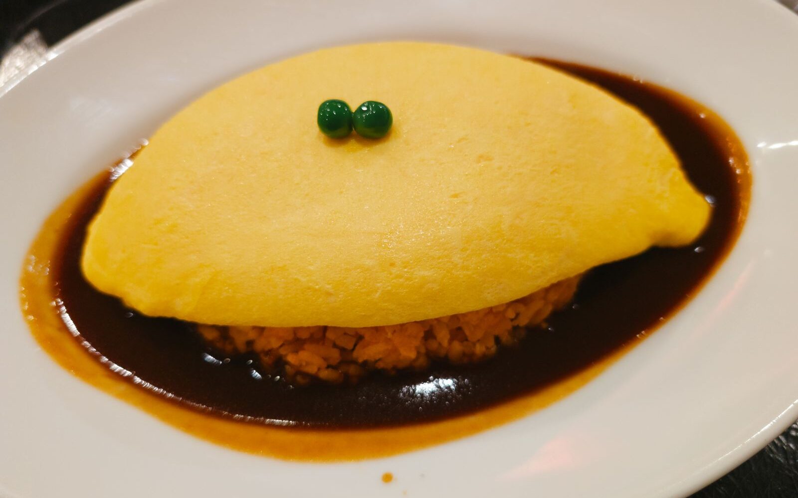 An omelette-wrapped rice dish, omurice, in a pool of brown sauce on a white plate, with two green peas on top