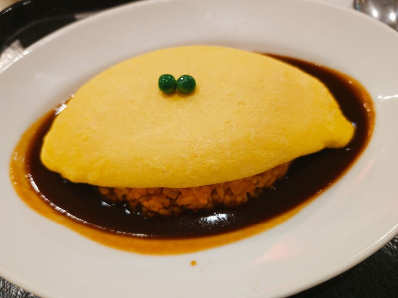 An omelette-wrapped rice dish, omurice, in a pool of brown sauce on a white plate, with two green peas on top
