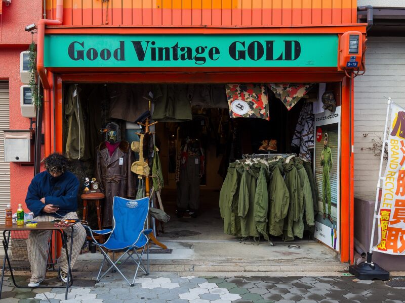 Open-fronted vintage clothing shop under a green Good Vintage GOLD sign, olive military parkas on a rail inside and a man seated at a table outside