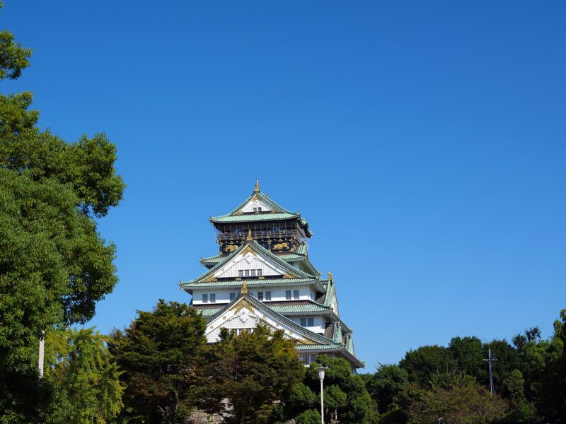 Osaka Castle's white keep rises above green park trees and stone walls beneath a clear blue sky.
