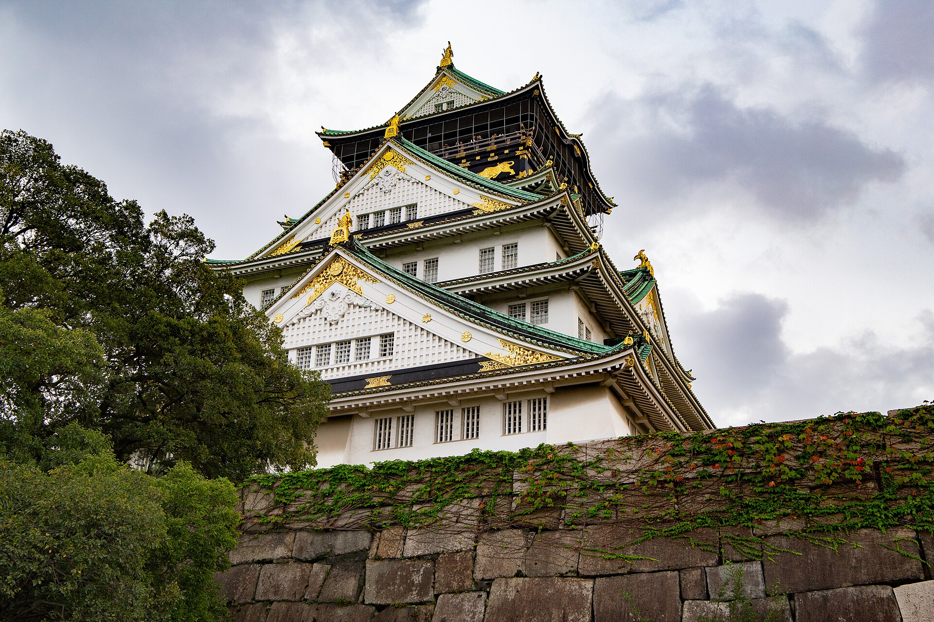Osaka Castle's white walls and gold-trimmed green roofs rise above an ivy-covered base of massive stone blocks.