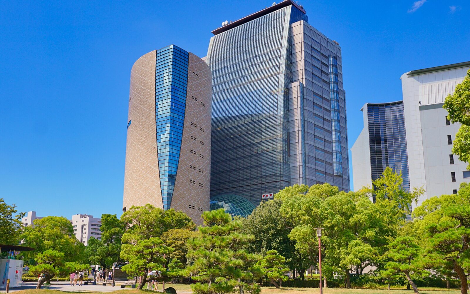 Osaka Museum of History's rounded tan tower stands beside the glass NHK Osaka building above green trees.
