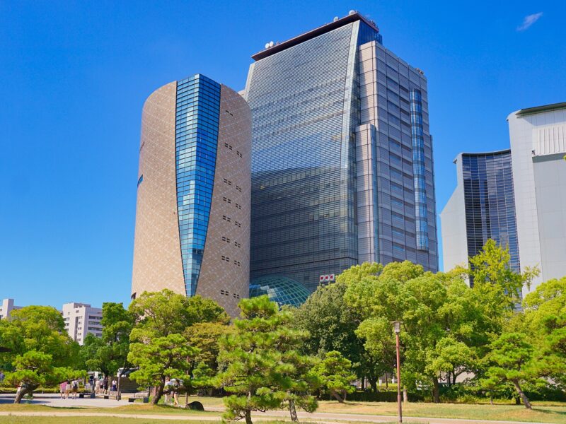 Osaka Museum of History's rounded tan tower stands beside the glass NHK Osaka building above green trees.