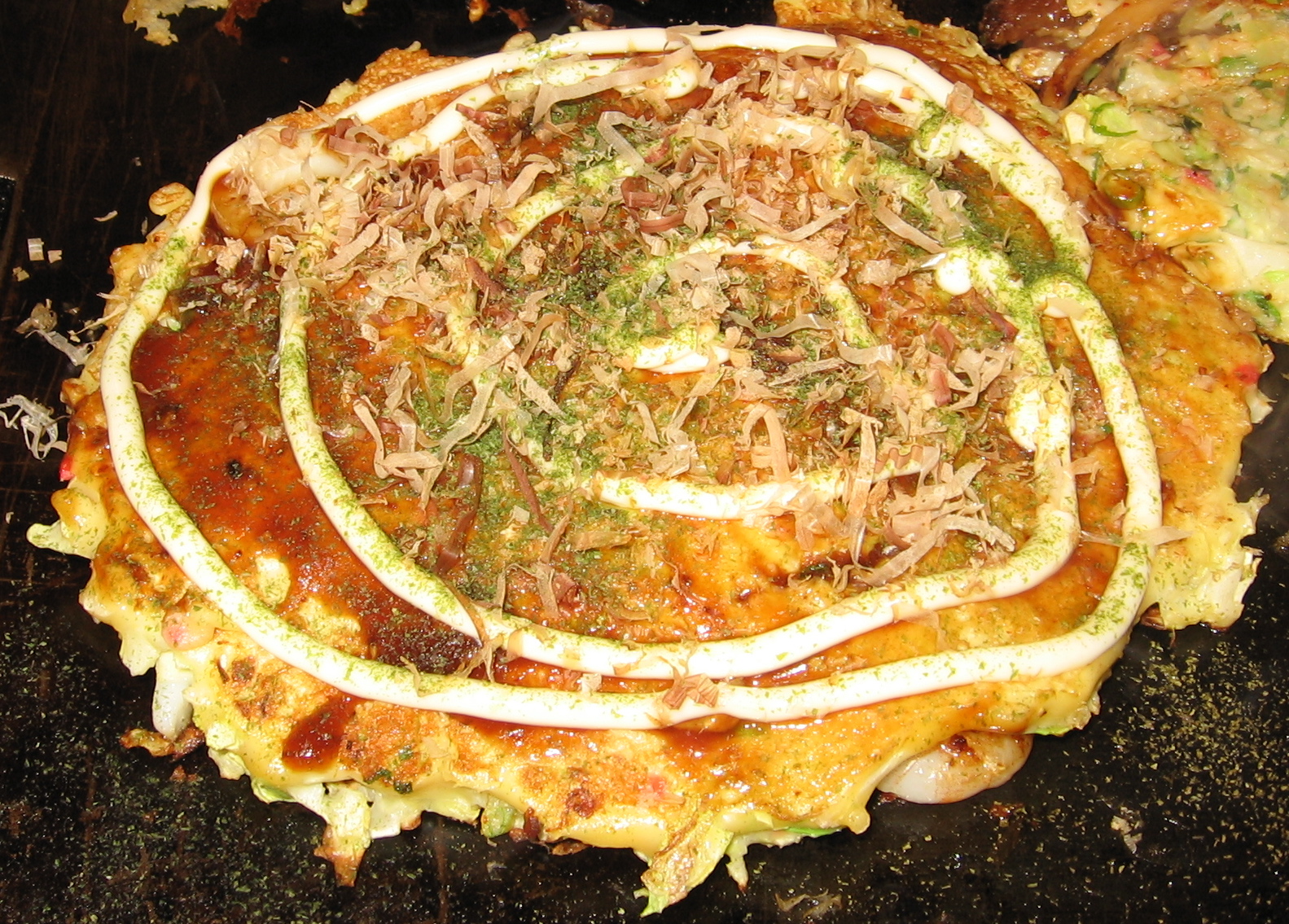 An Osaka okonomiyaki pancake sits on a dark griddle, topped with sauce, mayonnaise, green seasoning and bonito flakes.