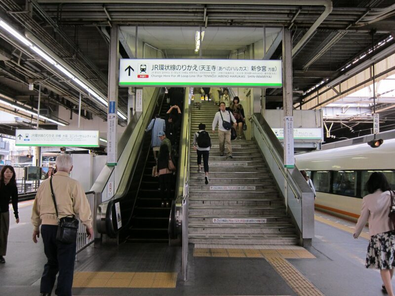 Passengers ride an escalator in single file beside a staircase and train platforms at Tsuruhashi Station in Osaka.