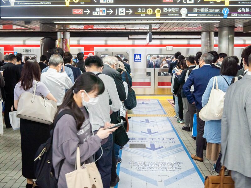 Passengers wait beside a red-striped train at Osaka Metro Umeda Station while a pedestrian checks a smartphone.