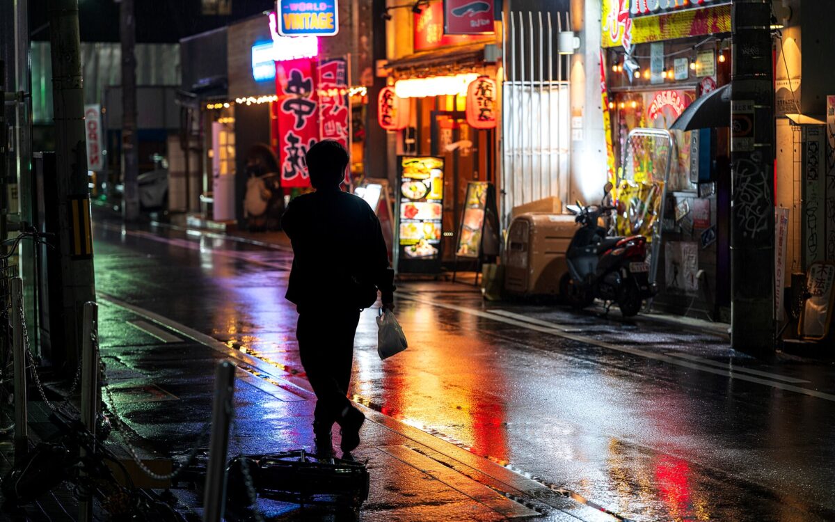 A pedestrian carrying a small bag walks along a wet Osaka street at night beneath colourful illuminated restaurant signs.