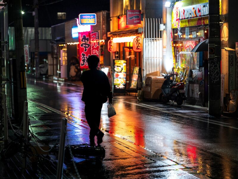 A pedestrian carrying a small bag walks along a wet Osaka street at night beneath colourful illuminated restaurant signs.