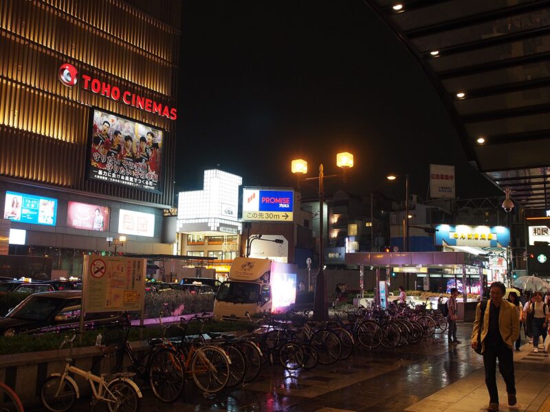 Pedestrians carry umbrellas past bicycles and glowing cinema signs on a wet evening in Namba, Osaka.