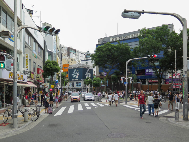 Pedestrians cross a shop-lined junction beside leafy Triangle Park in Osaka's Amerikamura district.