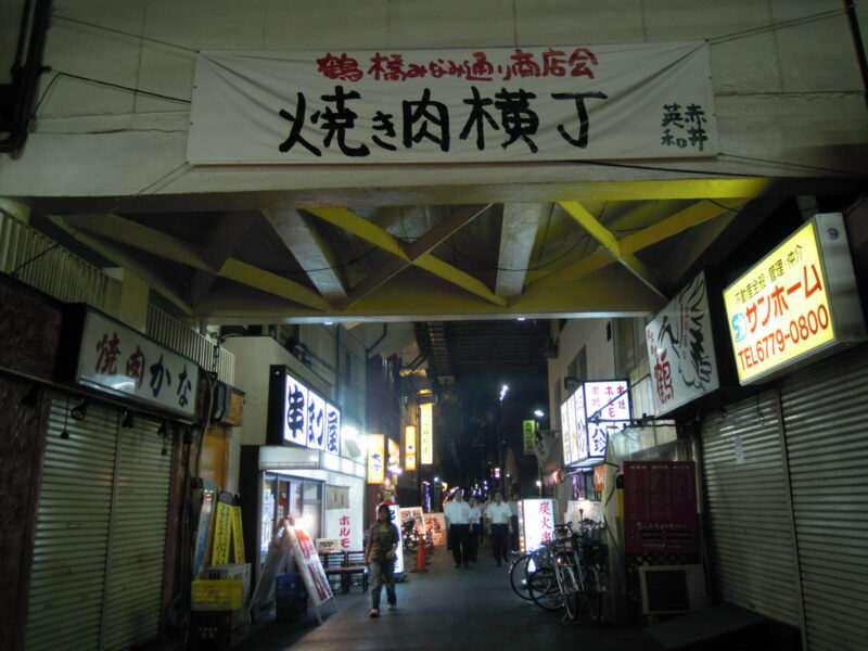 Pedestrians walk beneath the yakiniku alley banner among illuminated restaurant signs and bicycles in Tsuruhashi, Osaka.