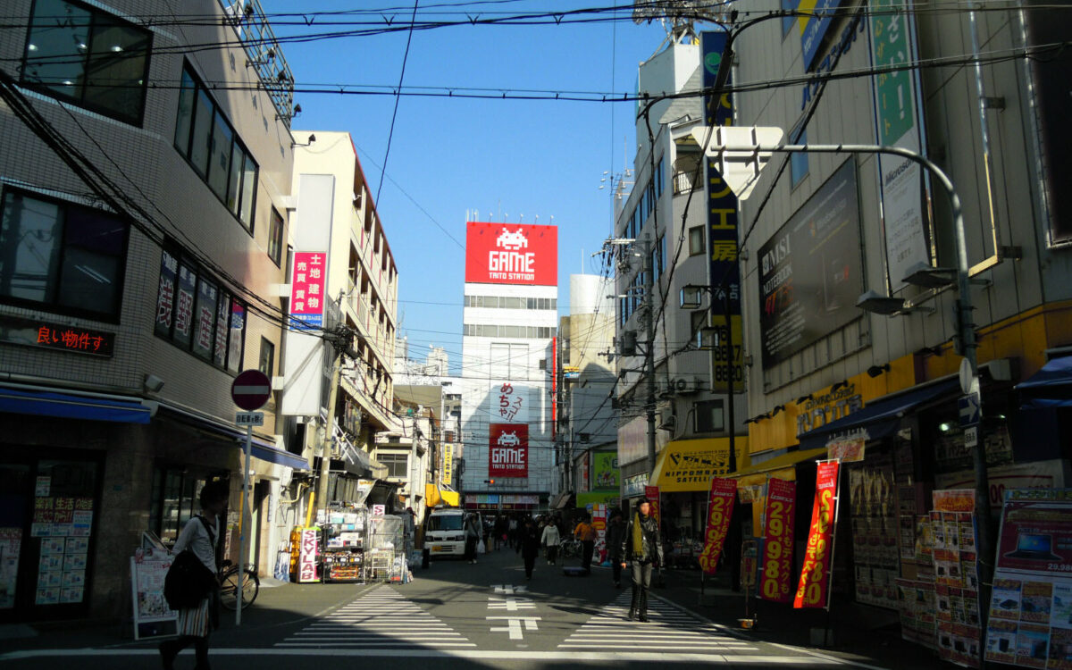 Pedestrians walk past electronics shops and a red Taito game sign on Otaku Road in Osaka’s Den Den Town.