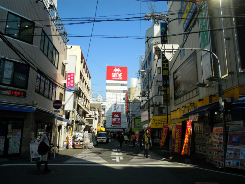 Pedestrians walk past electronics shops and a red Taito game sign on Otaku Road in Osaka’s Den Den Town.