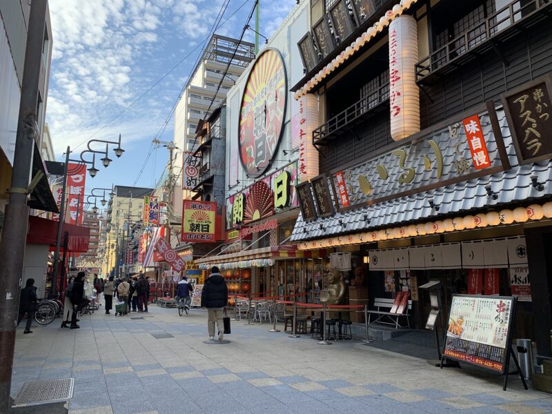 Pedestrians walk past lanterns, large restaurant signs and outdoor stools along Shinsekai Hondori shopping street in Osaka.