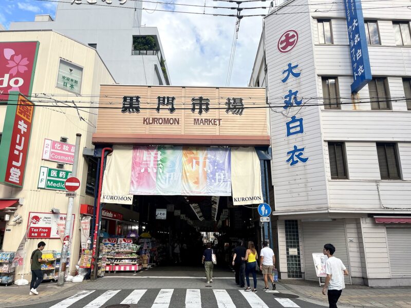 People enter Kuromon Market in Osaka beneath a wooden gateway sign and pale hanging curtains on a sunny day.