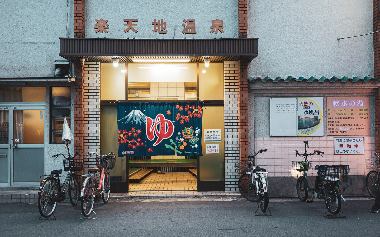 Rakutenchi Onsen's illuminated entrance in Osaka, with a Mount Fuji bathing curtain and bicycles parked outside the tiled facade.