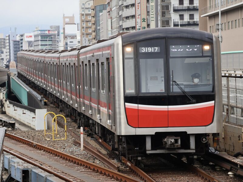 A red-and-silver Osaka Metro Midosuji Line train approaches Shin-Osaka on elevated tracks between city buildings.