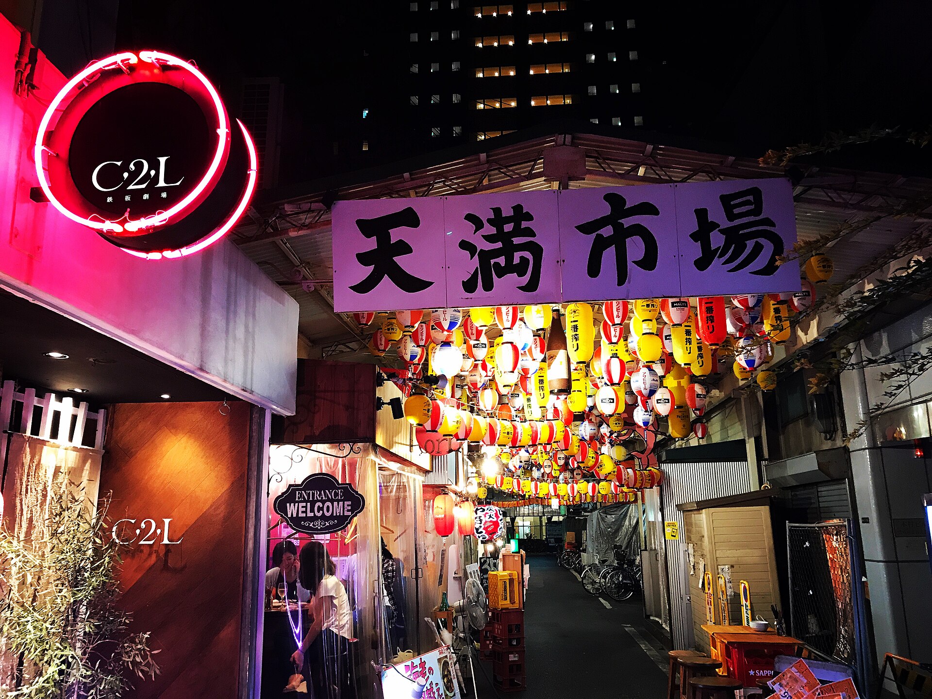 Red and yellow lanterns fill a narrow alley at Tenma market in Osaka, beside an open bar entrance and stools.