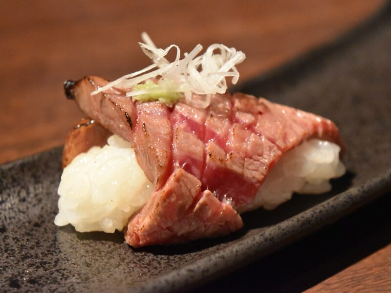 Seared Matsusaka beef slices cover two portions of sushi rice on a dark rectangular plate in Osaka.