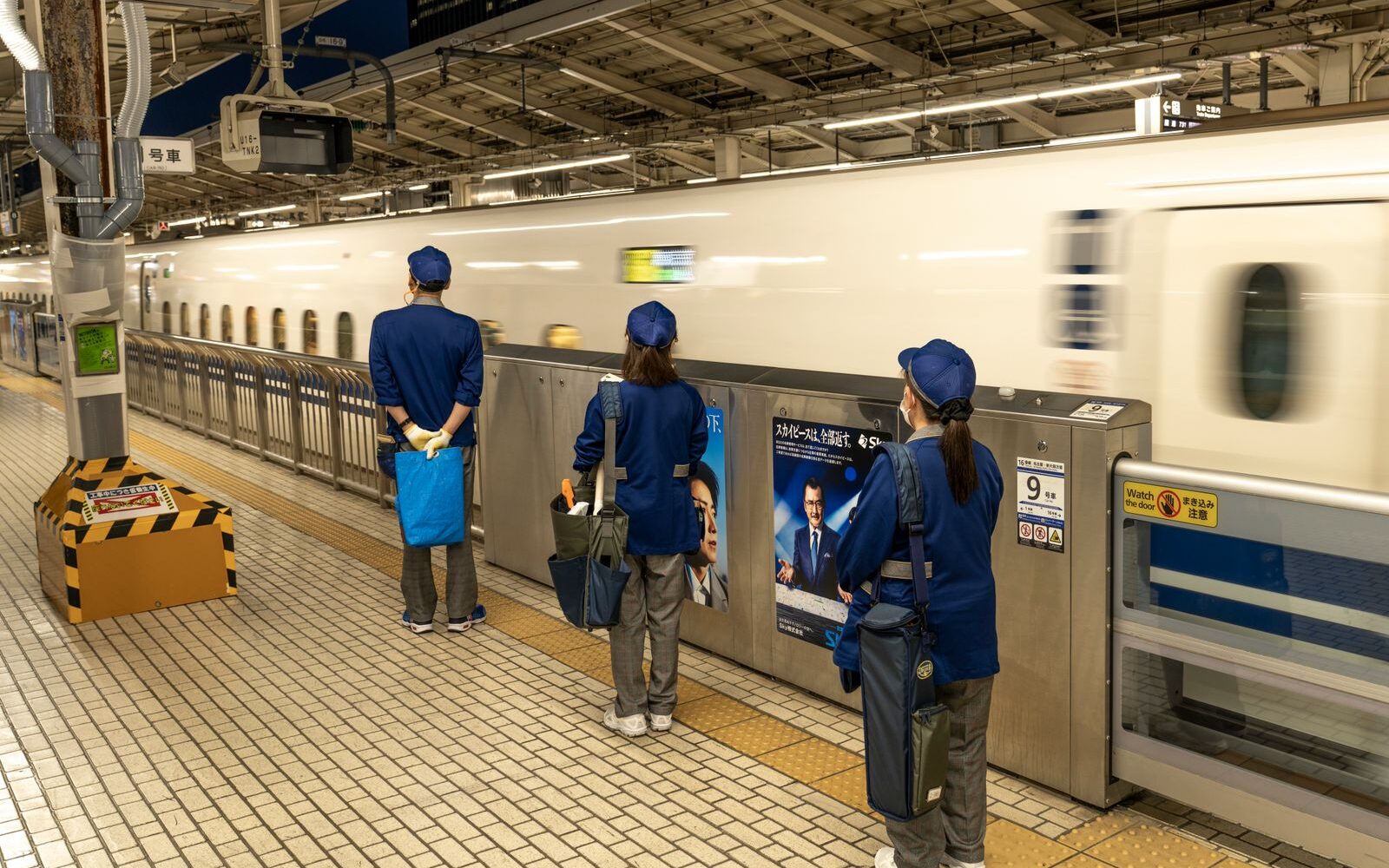 A cleaning crew in blue uniforms waits at the platform doors as a white and blue Tokaido Shinkansen pulls into the station