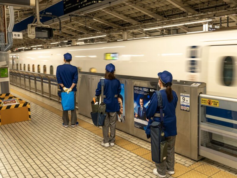 A cleaning crew in blue uniforms waits at the platform doors as a white and blue Tokaido Shinkansen pulls into the station