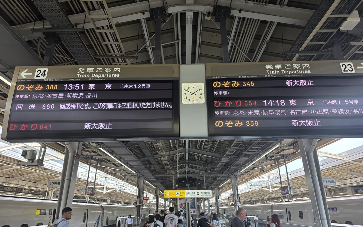 Shinkansen trains flank a busy Shin-Osaka platform beneath departure boards for tracks 23 and 24, with passengers carrying luggage.