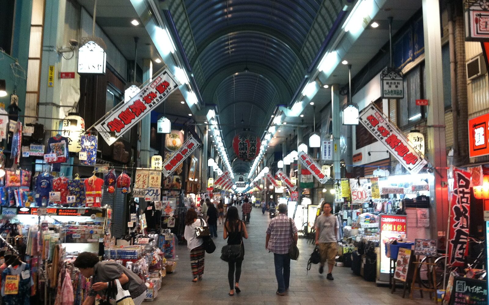 Shoppers browse merchandise and walk between clothing displays beneath the covered roof of Tenjinbashisuji Shopping Street in Osaka.