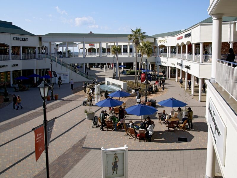 Shoppers cross the palm-lined courtyard at Rinku Premium Outlets, with blue parasols and two levels of shops.