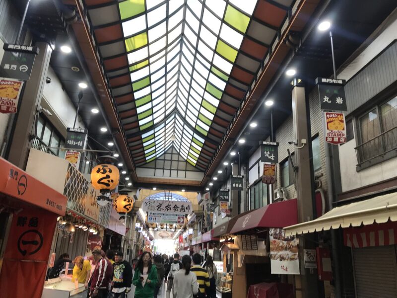 Shoppers walk between food stalls beneath the multicolored roof panels of Kuromon Market's covered arcade in Osaka.