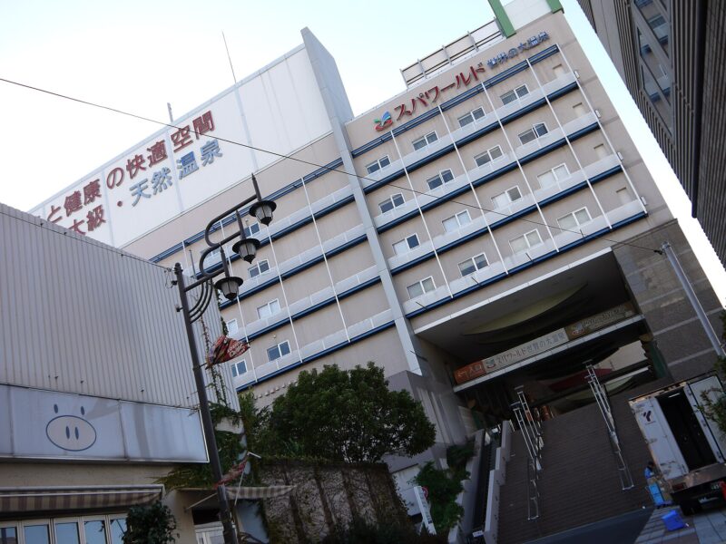 Spa World's tall facade and broad entrance stairs in Osaka, with Japanese lettering above the street and trees.