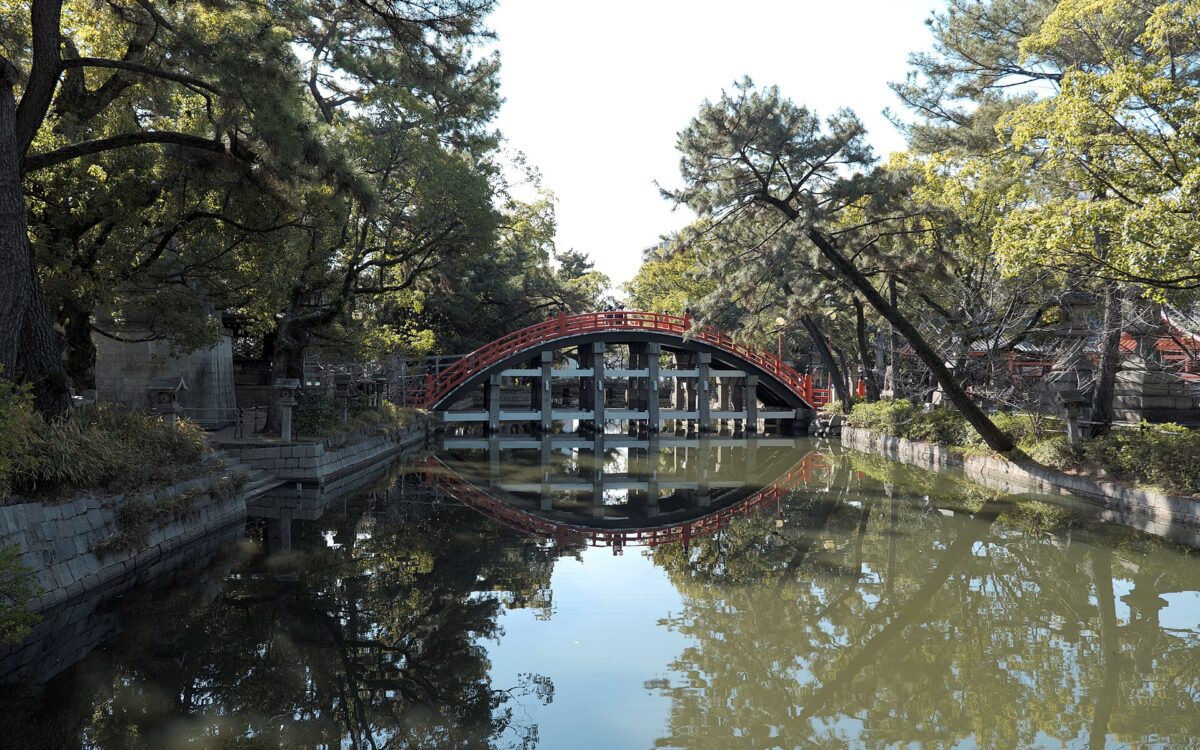 Sumiyoshi Taisha’s vermilion Sorihashi bridge arches over a tree-lined pond in Osaka, with its railings and supports reflected in the water.