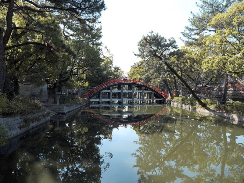 Sumiyoshi Taisha’s vermilion Sorihashi bridge arches over a tree-lined pond in Osaka, with its railings and supports reflected in the water.
