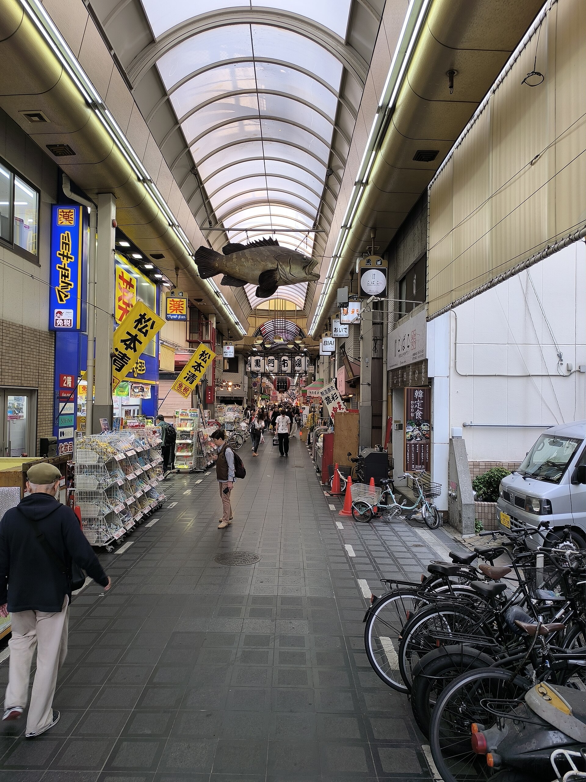 A suspended fish overlooks Kuromon Market's covered aisle in Osaka, with shops, passing pedestrians and bicycles along the sides.
