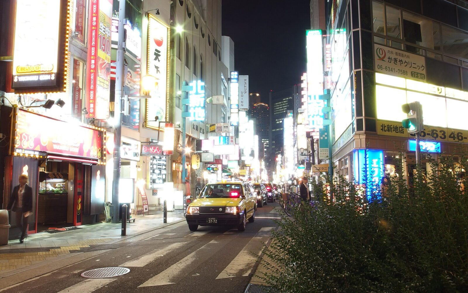 A taxi approaches along a Kitashinchi street in Osaka beneath rows of illuminated restaurant and bar signs.