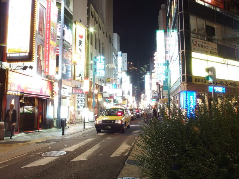 A taxi approaches along a Kitashinchi street in Osaka beneath rows of illuminated restaurant and bar signs.
