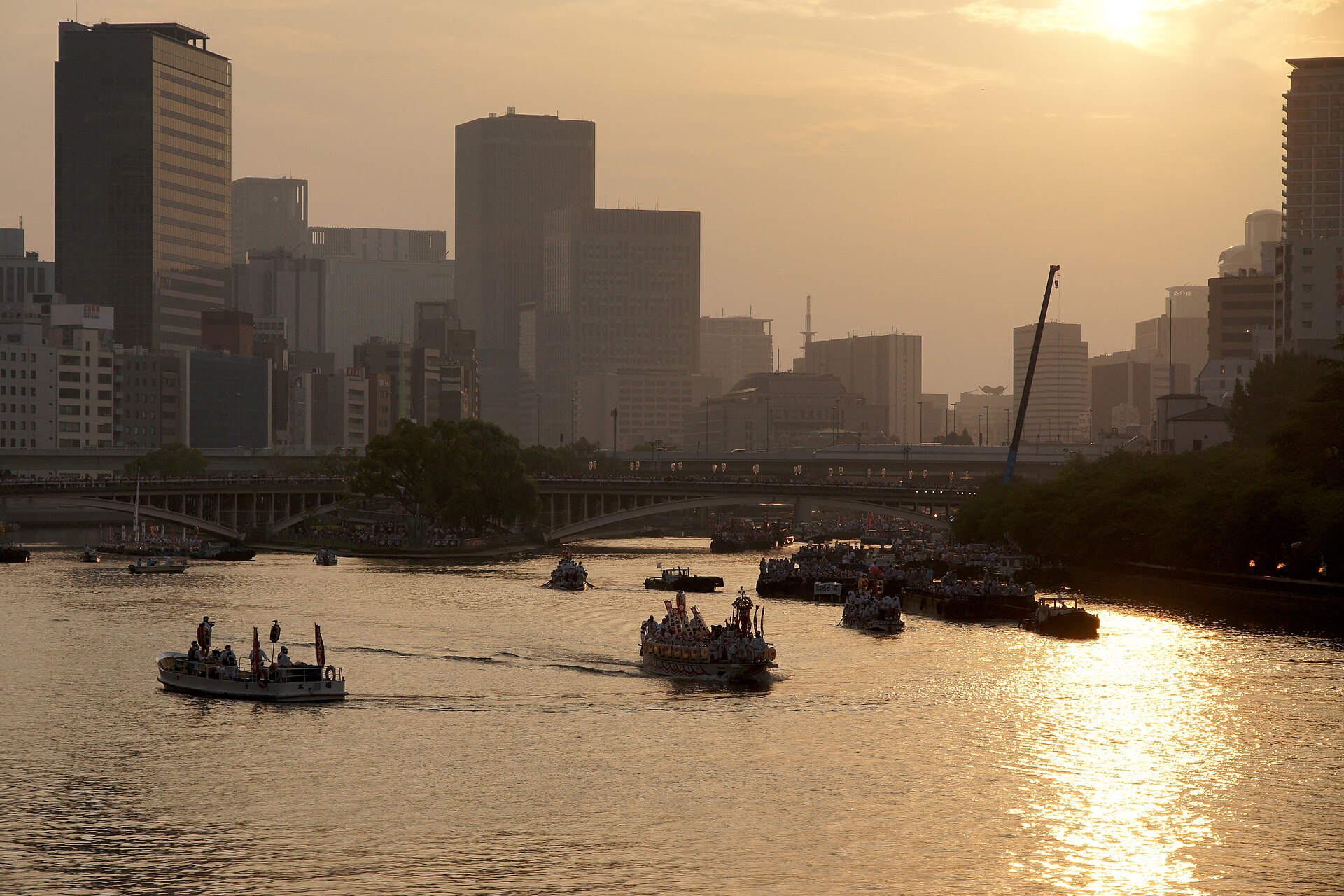 Tenjin Matsuri boats cross the sunlit Okawa River beneath bridges and tall Osaka buildings on a summer evening.