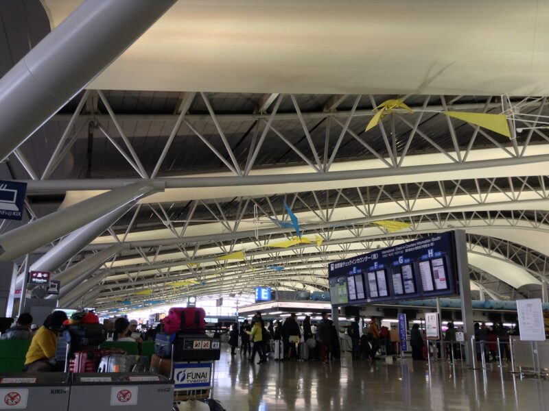 Travelers and luggage trolleys beneath the curved roof of Kansai Airport Terminal 1, beside international check-in information boards.