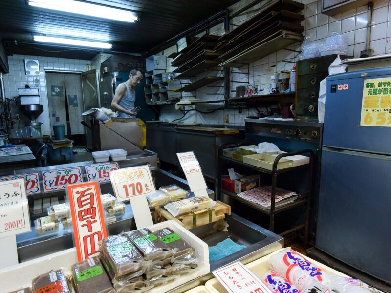 Trays of fresh tofu and sesame tofu with handwritten price cards at the counter of a small tofu maker's shop in Taisho, Osaka