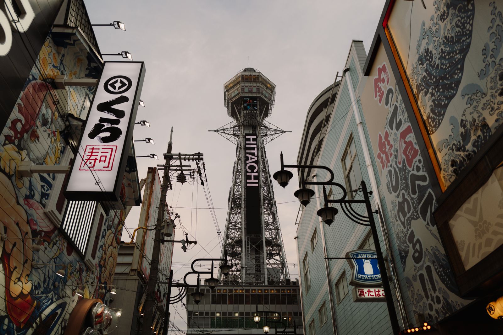 Tsutenkaku Tower with its vertical HITACHI sign, seen between buildings painted with large murals