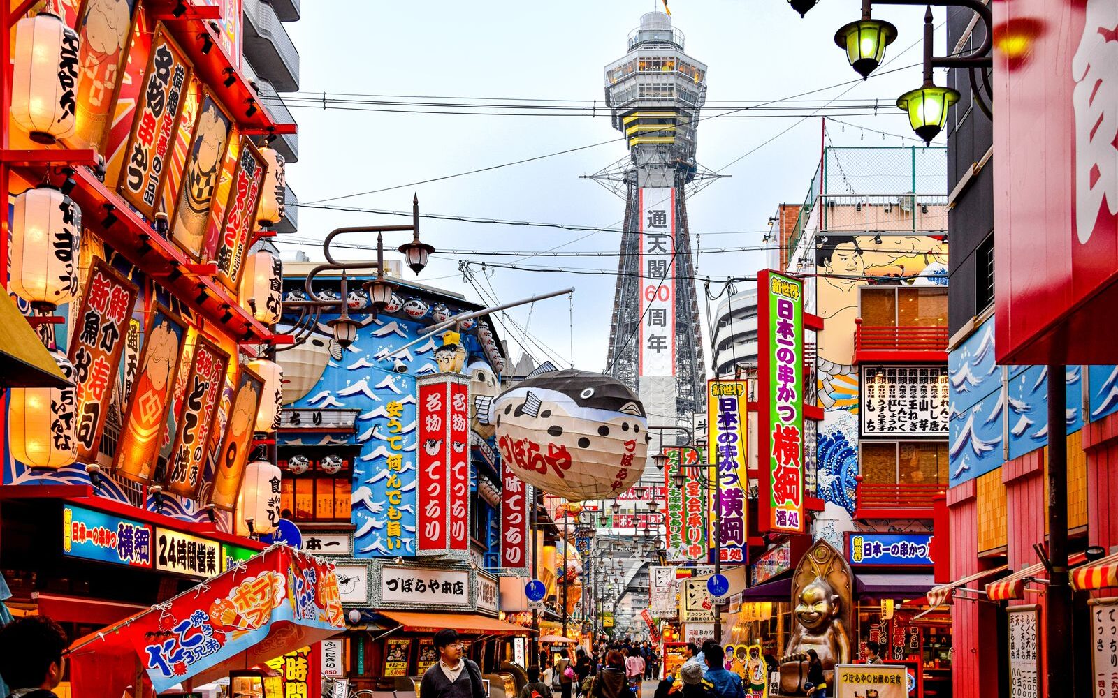 Tsutenkaku Tower at the end of a busy Shinsekai street lined with lanterns, neon signs and a giant pufferfish lantern