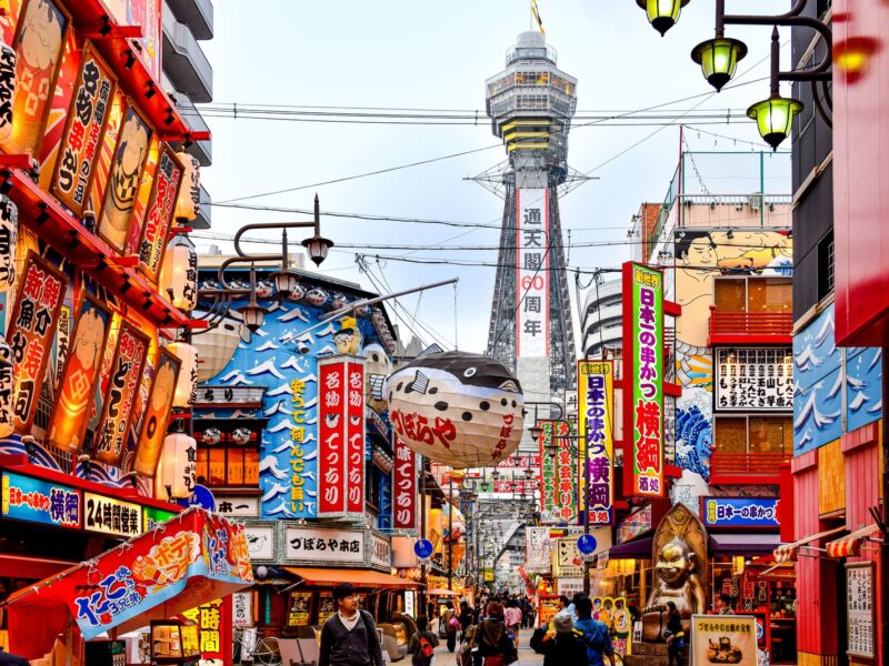 Tsutenkaku Tower at the end of a busy Shinsekai street lined with lanterns, neon signs and a giant pufferfish lantern