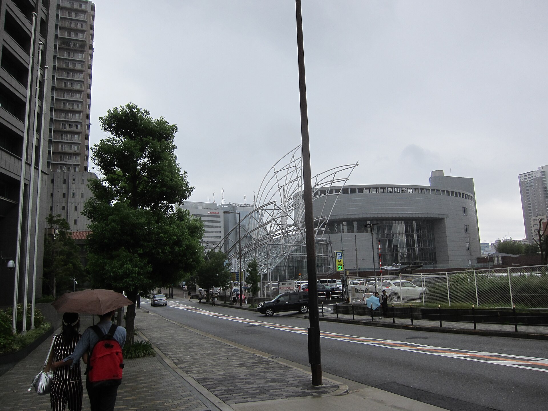 Two pedestrians share an umbrella on a wet Osaka street beside the National Museum of Art's metal framework.