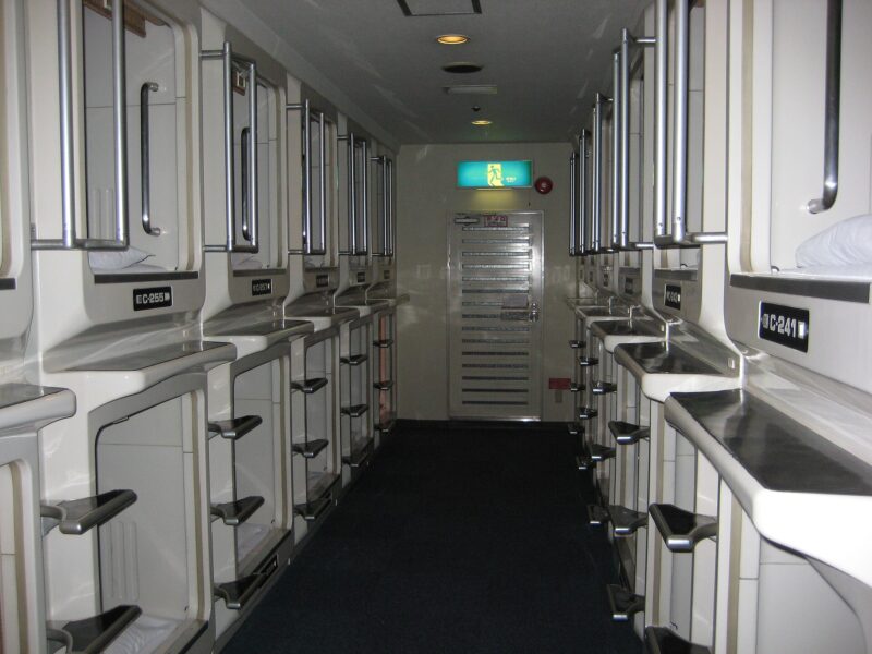 Two tiers of white sleeping pods line a narrow aisle inside an Osaka capsule hotel, with metal steps and handrails.